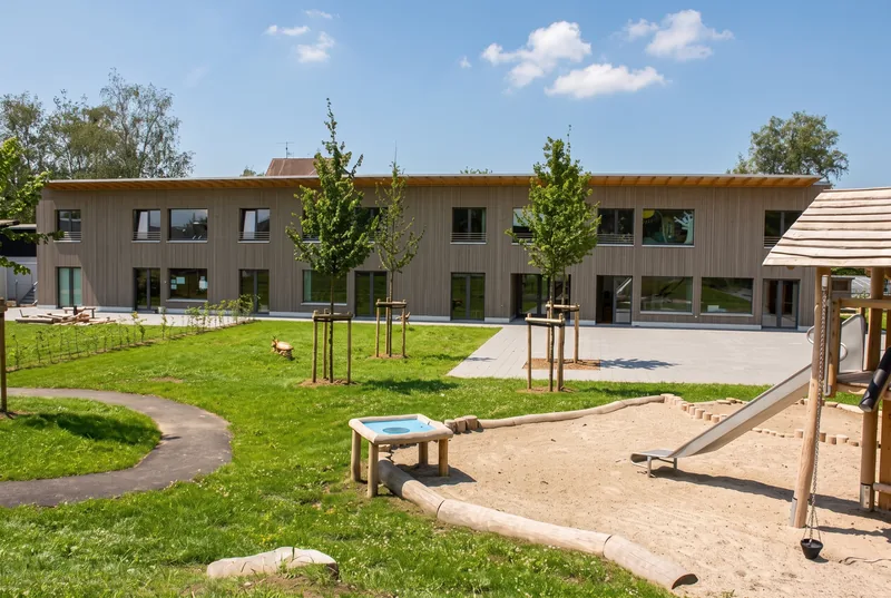 Garten der Kita mit Rasen, Sandflächen und Spielgeräten vor dem langgestreckten Gebäude